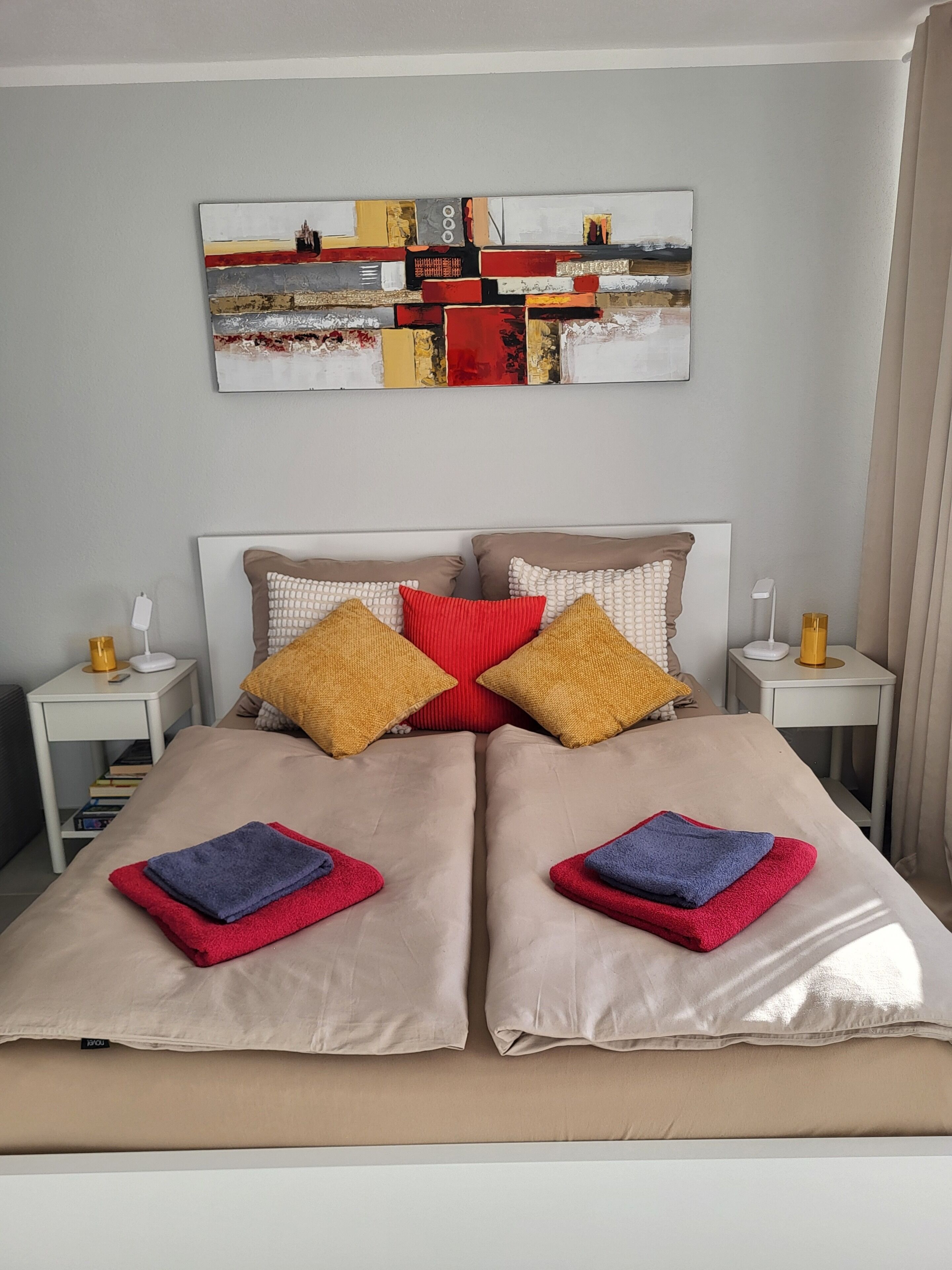Un letto matrimoniale con lenzuola beige, due cuscini rossi e due gialli, e due asciugamani piegati. Sopra il letto c'è un quadro e su ciascun lato del letto un comodino con una lampada e un telefono.