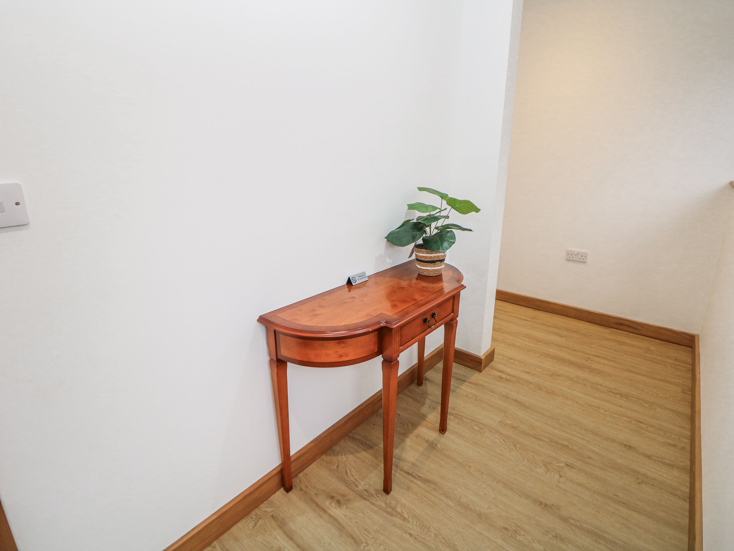 Una consola de madera con una planta, ubicada frente a una pared blanca en una habitación con piso de madera.