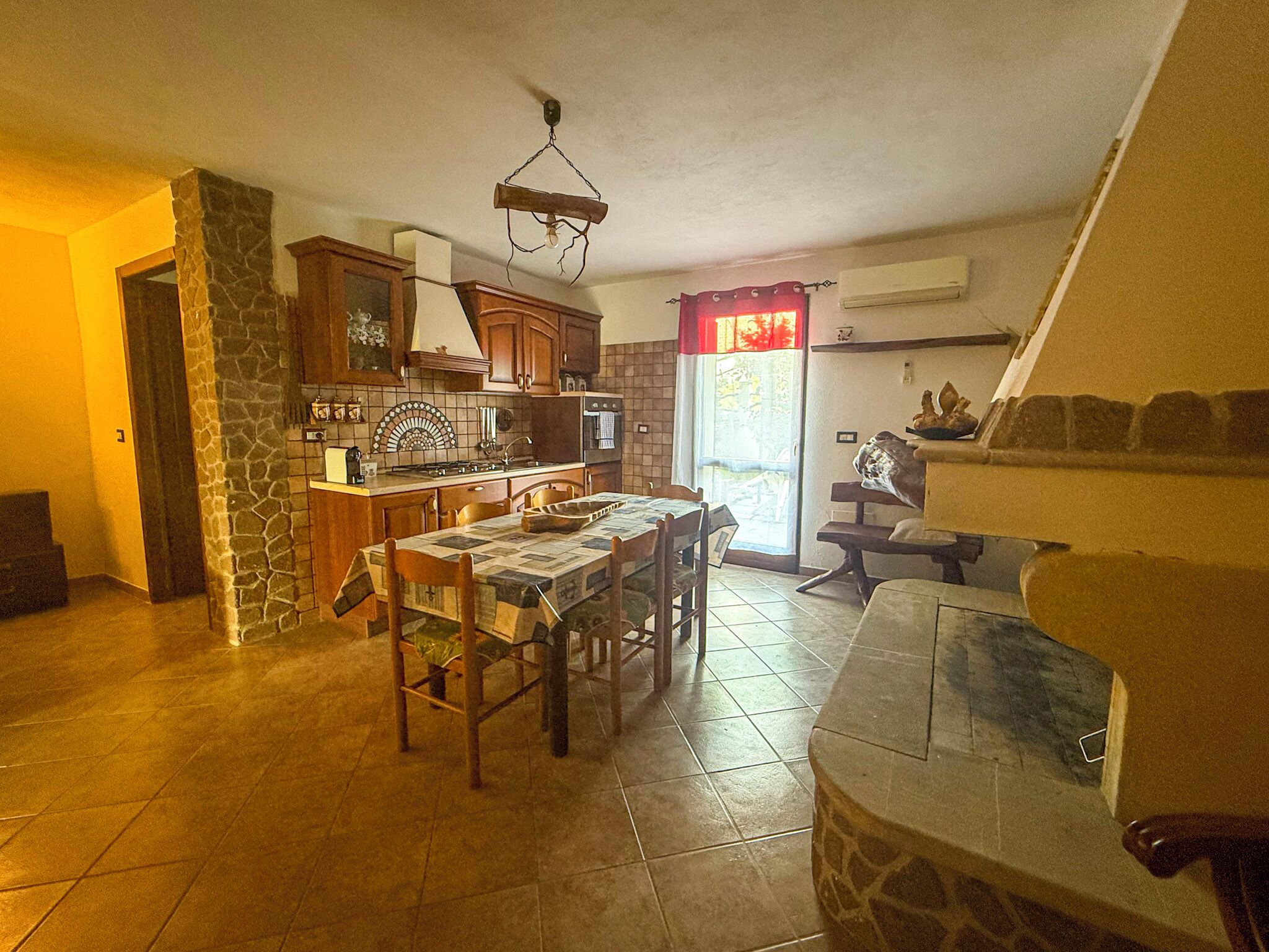 Cuina amb paret de pedra, armaris de fusta, sòl de rajola i zona de dinar amb taula i cadires.