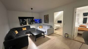 Un salon moderne avec un canapé noir, un canapé blanc et une table basse. Un téléviseur à écran plat est fixé au mur, et un tableau est accroché au-dessus du canapé blanc.