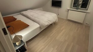 A small, well-lit room with a single bed, a bedside table with a lamp, a TV mounted on the wall, and a window with a view.