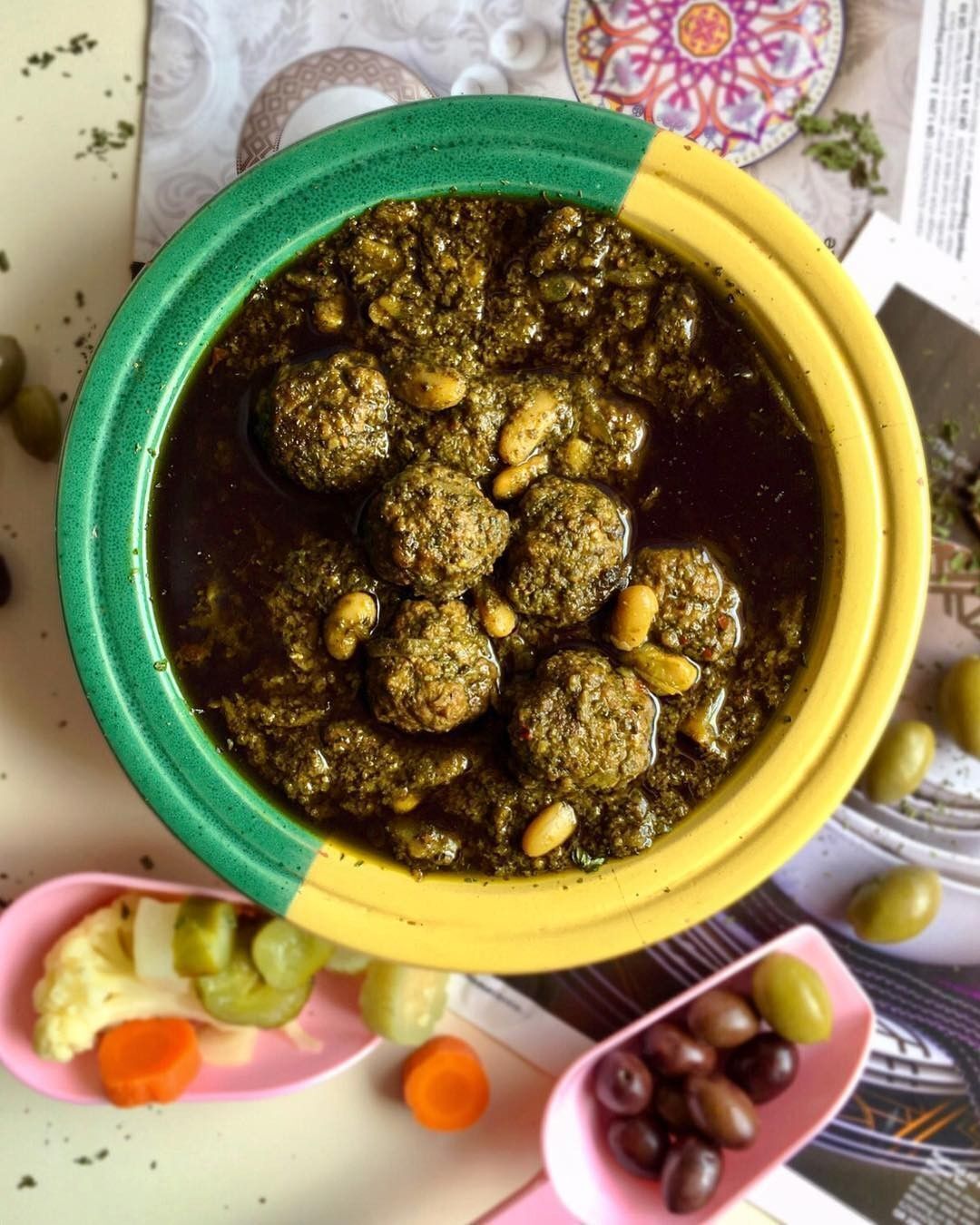 Un tazón de salsa verde con albóndigas y aceitunas, rodeado de verduras y aceitunas sobre una mesa.