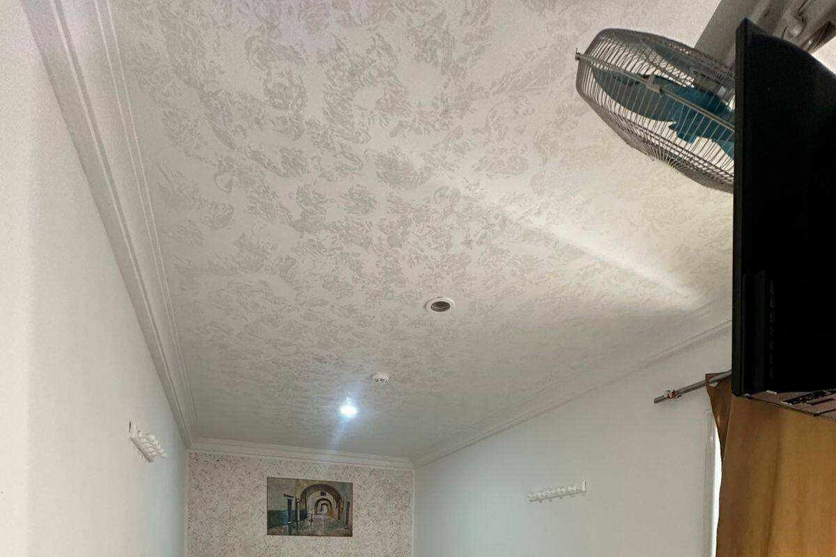 Une petite pièce bien éclairée, avec deux lits simples, un radiateur, un ventilateur de plafond et un tableau accroché au mur.