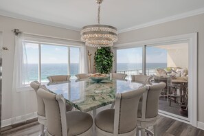 A dining area with a glass table, beige chairs, a chandelier, and a view of the ocean. - Luxury Penthouse at Regency Towers | 4 Bedrooms | Gulf Views (Panama City Beach)