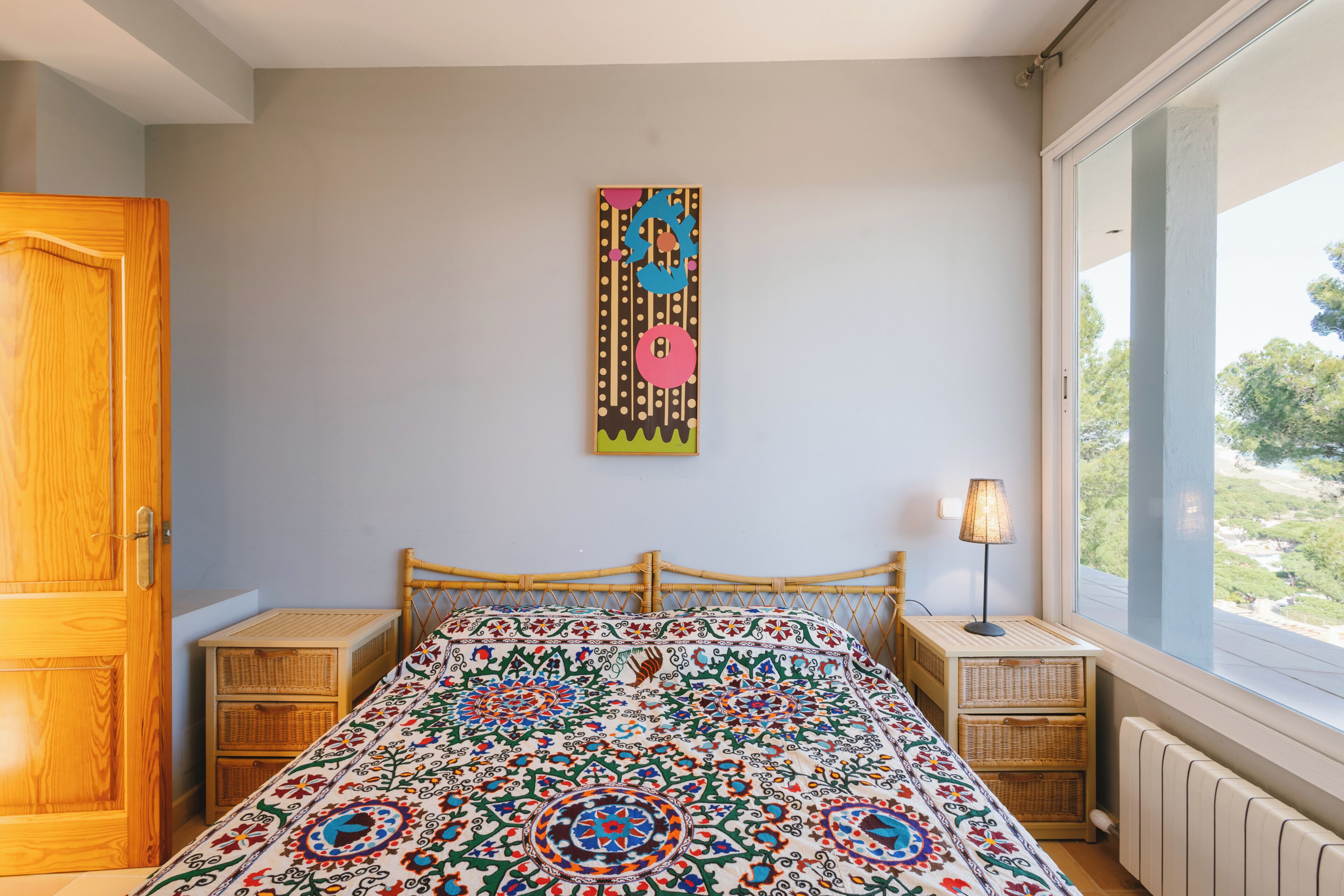 Ein Schlafzimmer mit einem hölzernen Bett, einem farbenfrohen Wandteppich, einem Nachttisch mit Lampe und einem Fenster mit Blick.