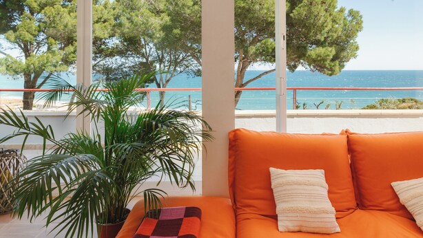 Een balkon met uitzicht op zee en bomen, een plant in pot, en een oranje bank met kussens.