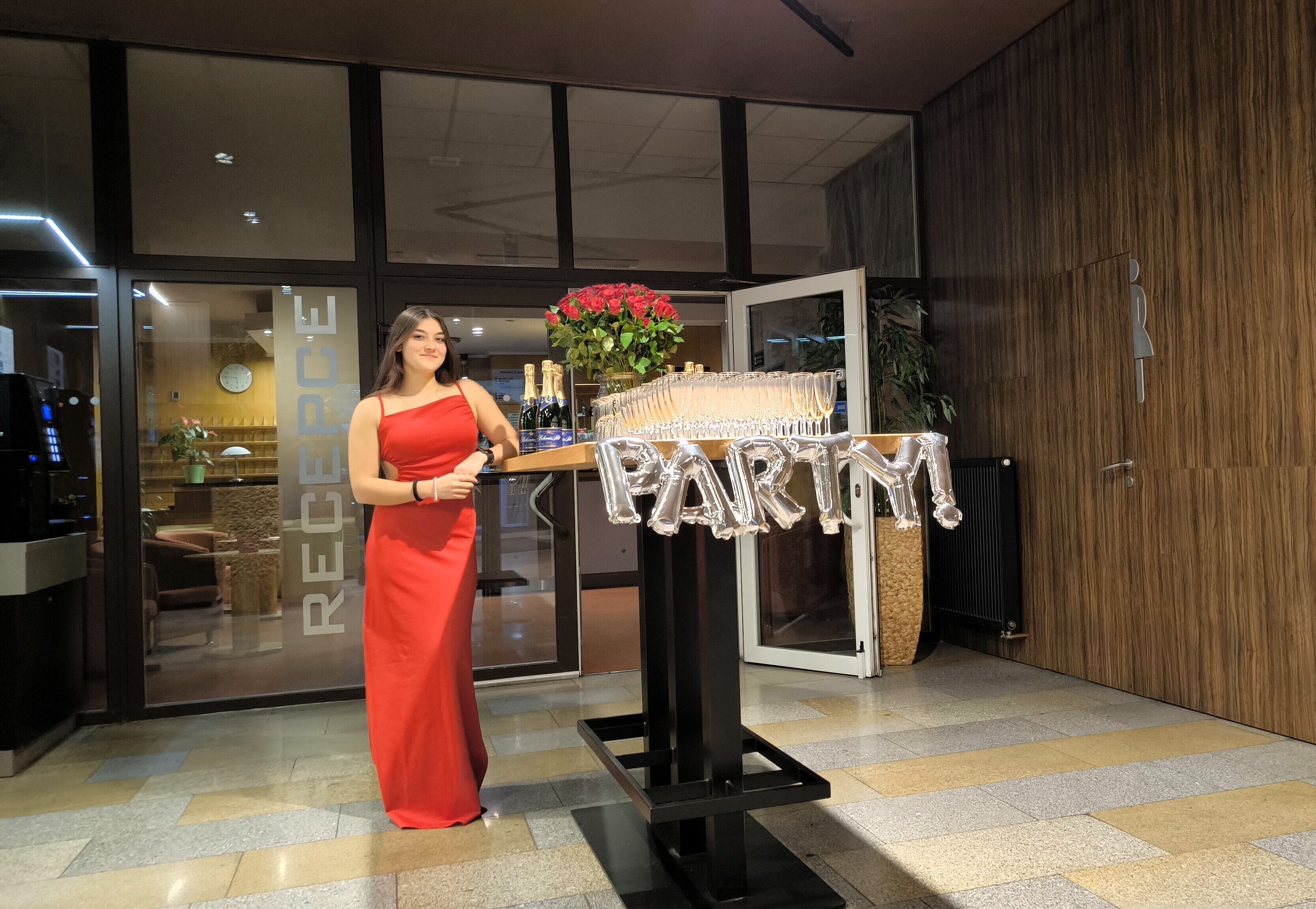 En kvinna i en röd klänning står framför en receptionsdisk med en skylt som visar ordet ”RECEPTION”, en blomsteruppsättning och champagneflaskor.