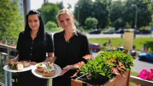 Two individuals holding trays with food items on a balcony with a view of a parking area and greenery.