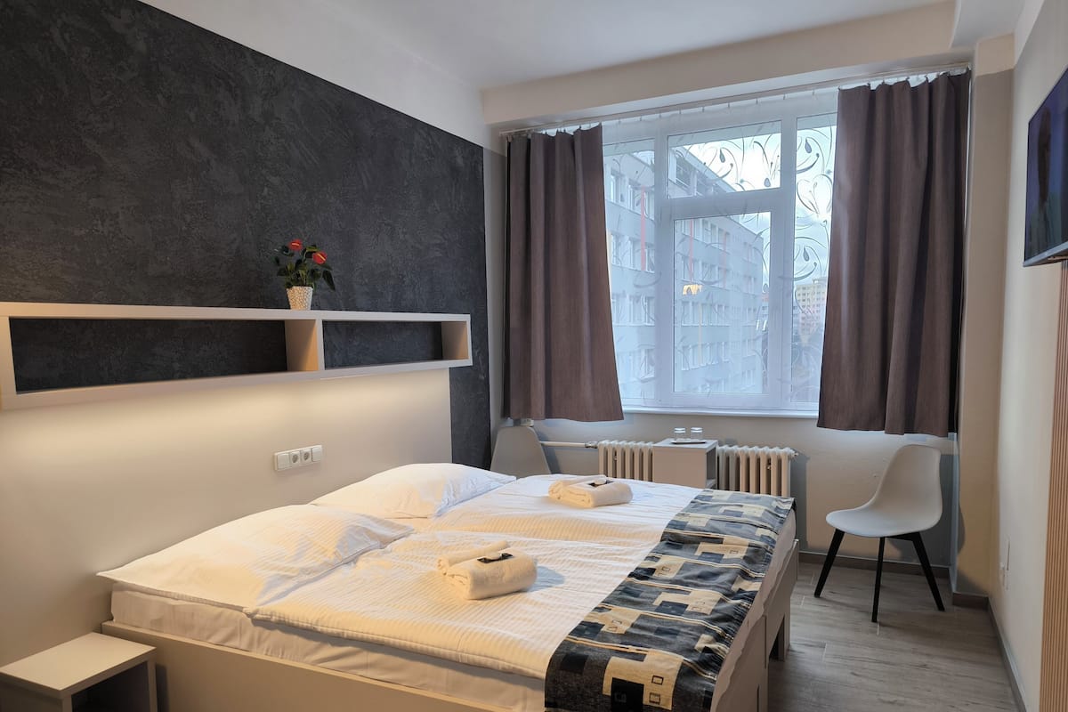 Habitación de hotel moderna con cama, mesita de noche, silla, ventana con cortinas, radiador y estante con una planta.