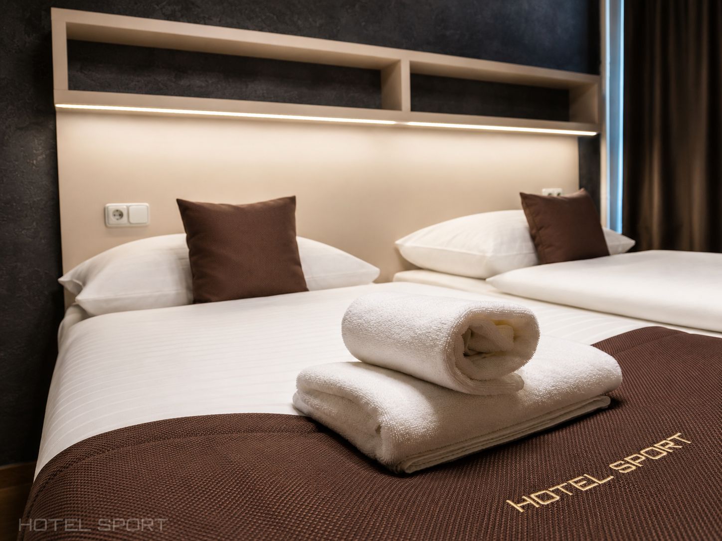 A hotel room with two beds, each with a brown pillow, a white blanket, and a folded towel on the bed. There is a shelf above the beds and a wall socket.