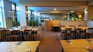 A restaurant with wooden tables and chairs, large windows, and indoor plants.