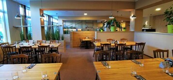 Un restaurante con mesas y sillas de madera, ventanas amplias y plantas de interior.