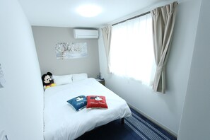 Habitación de hotel con cama, un peluche de Mickey Mouse, dos almohadas con motivos de Mickey Mouse, aire acondicionado de pared y una ventana con cortinas.