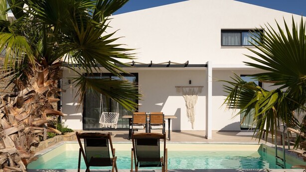 Uma área de piscina moderna, com duas espreguiçadeiras de madeira, um edifício branco com grandes janelas e um detalhe decorativo de macramé suspenso.