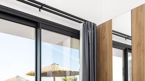Quarto de hotel moderno com uma grande porta de correr de vidro que dá para uma área de piscina, mobiliado com espreguiçadeiras e um guarda-sol de palha.