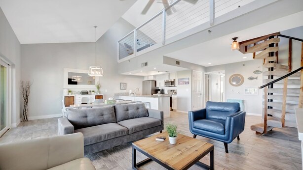 A spacious living area with a gray sofa, a blue armchair, and a wooden coffee table. A kitchen is visible in the background with white cabinets and a modern design. - Sleep 8 next to the Beach! Balcony & Heated Pool (Indian Shores)
