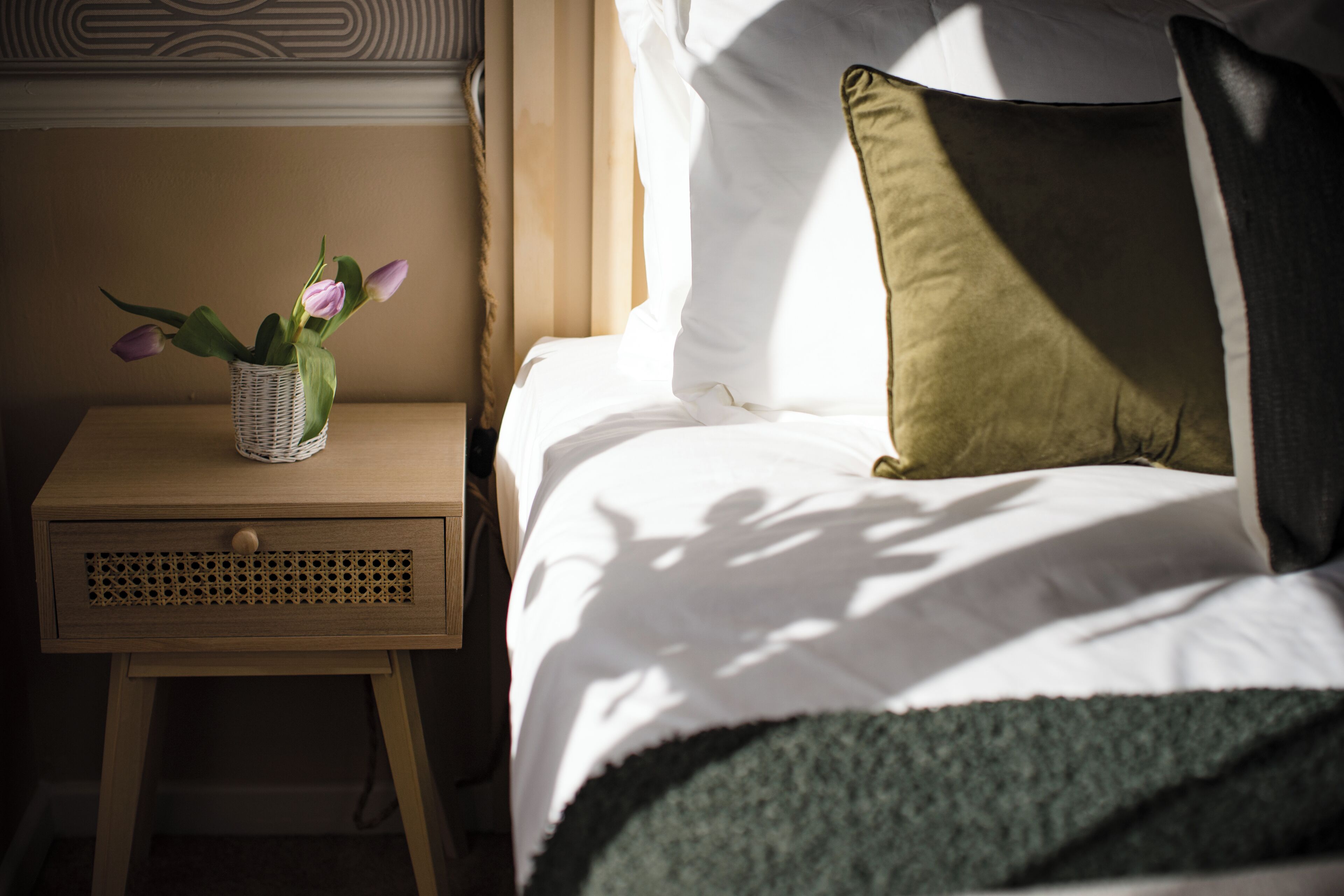 A neatly made bed with a green throw blanket, a pillow, and a small bedside table with a wicker basket containing flowers.