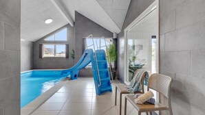 An indoor swimming pool with a blue slide, a seating area with a chair, and a view of the outside through large windows. - VillaVilla 541 - Bjerregård, Vestjylland (Hvide Sande)