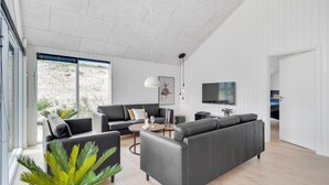 A modern living room with a large grey sectional sofa, a coffee table, and a TV mounted on the wall. There is a window with a view of a rocky landscape. - VillaVilla 541 - Bjerregård, Vestjylland (Hvide Sande)