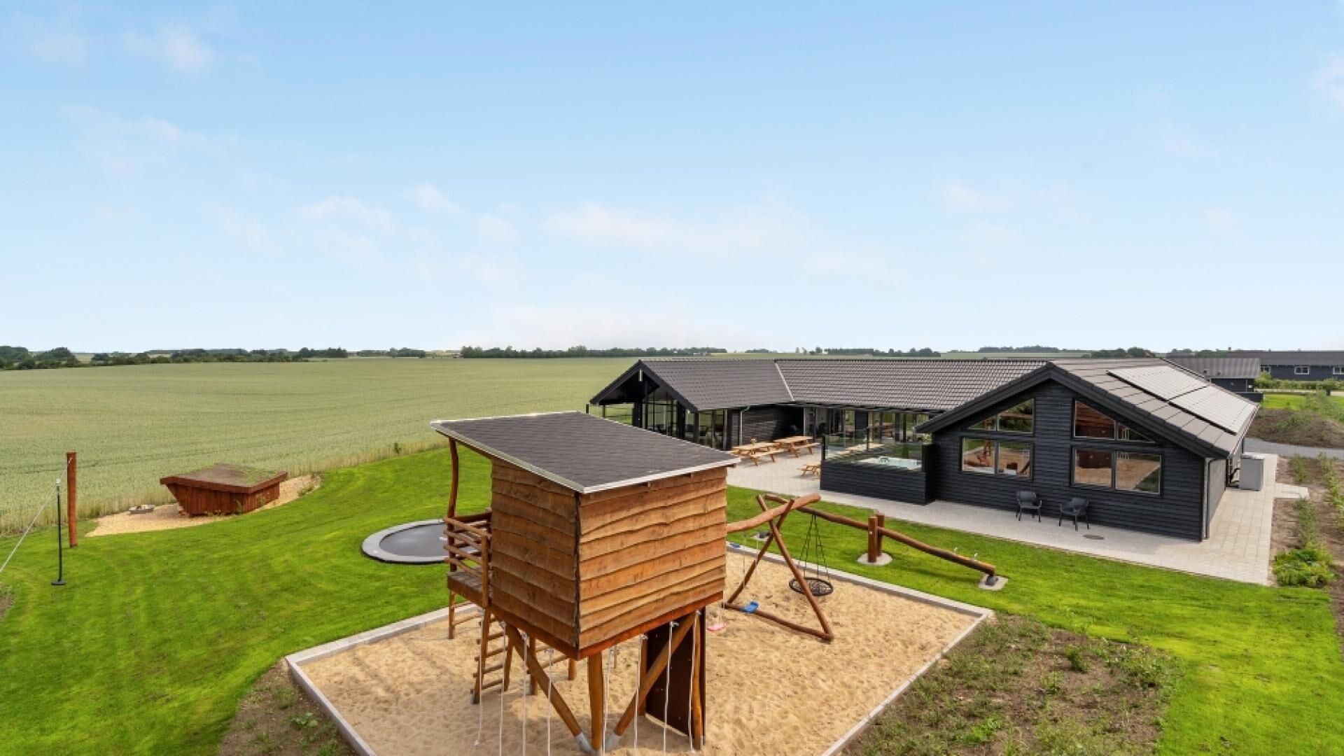 A modern cabin with a large deck and a swing set, surrounded by a grassy field and a large open area.