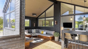 Un espace de vie extérieur moderne doté d’une terrasse en bois, d’un coin salon intégré et d’un bar avec des tabourets.
