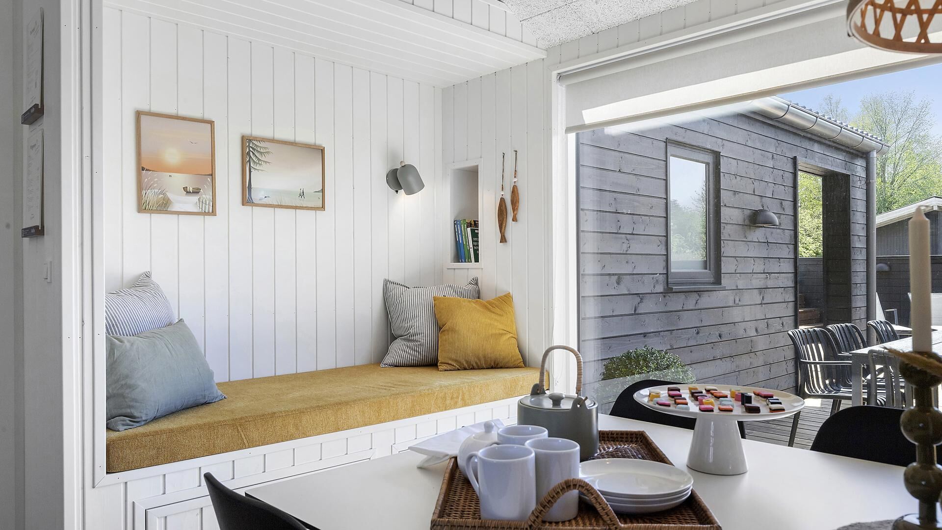 Une pièce chaleureuse avec un banc blanc, une table sur laquelle se trouve un plateau rempli d’objets, et une vue sur l’extérieur.