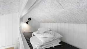 Une petite pièce aux murs recouverts de panneaux blancs, avec un plafond en pente, un lit simple avec du linge de lit blanc et une lampe de bureau noire.