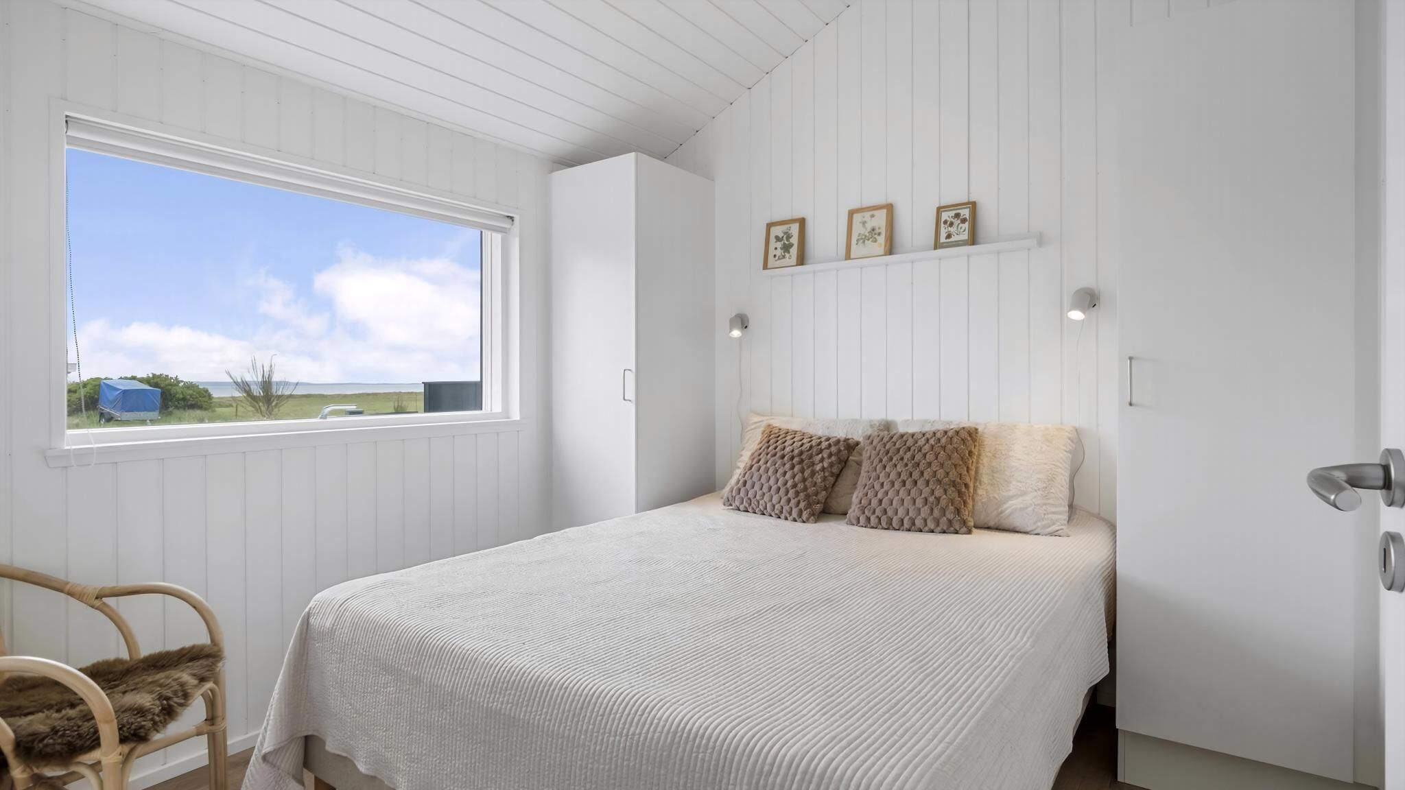Quarto com cama, uma cadeira, janela com vista e quadros nas paredes.