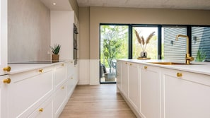 Eine moderne Küche mit weißen Schränken und goldenen Griffen, einem schwarzen Herd und einem großen Fenster mit Blick auf Grünflächen.