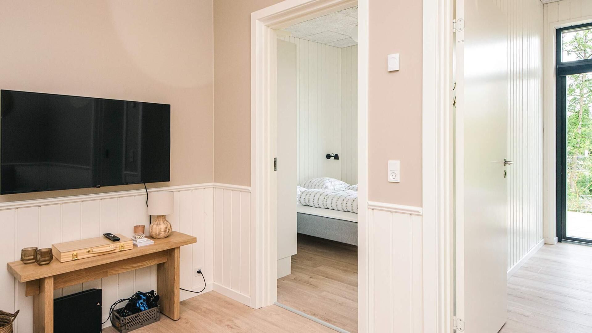 Una stanza con una televisione montata a parete, una scrivania in legno con una lampada e un letto con testiera visibile attraverso una porta aperta.