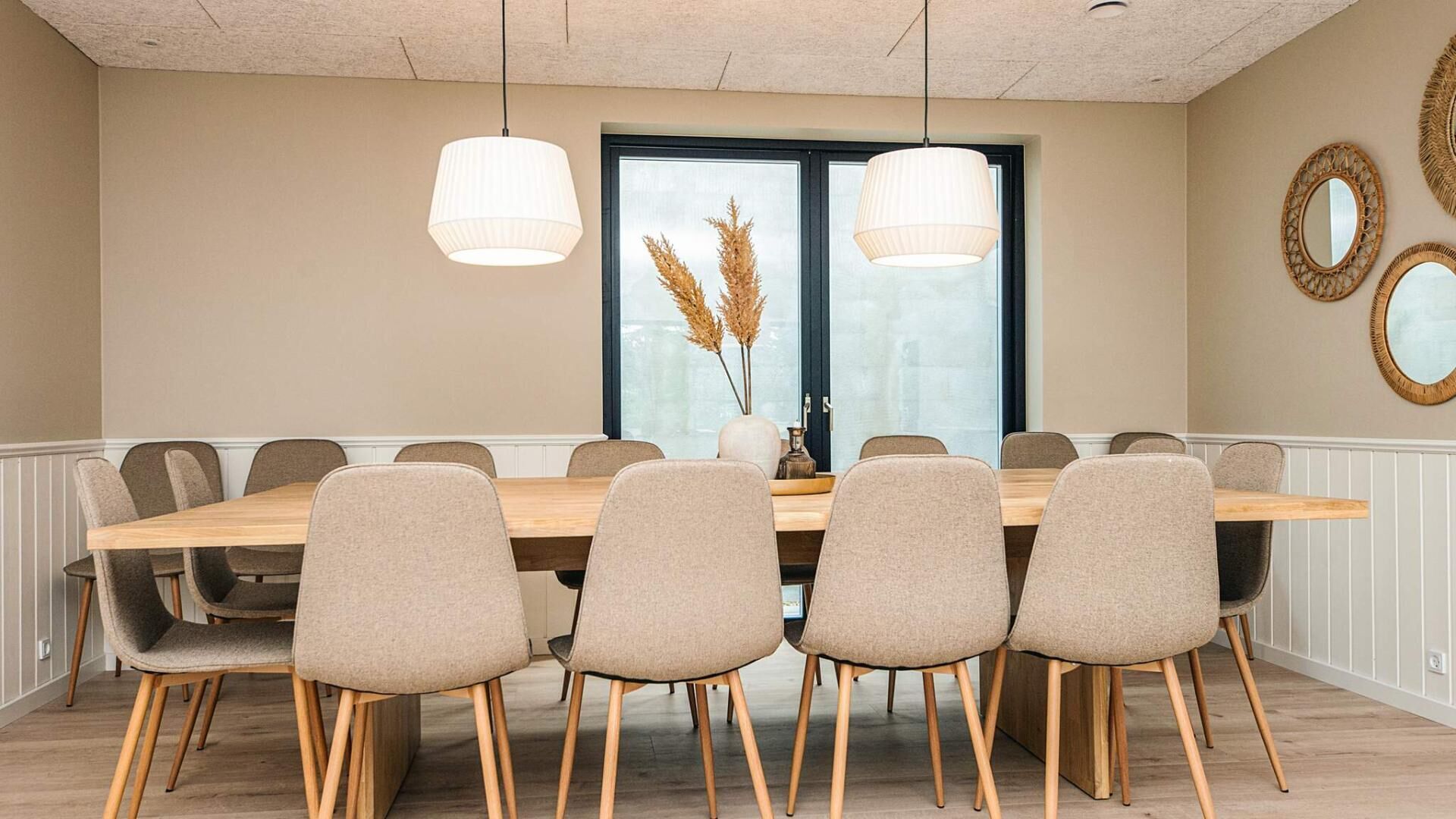 Une salle à manger avec une longue table en bois, six chaises rembourrées, deux lampes suspendues et une fenêtre agrémentée de plantes décoratives.