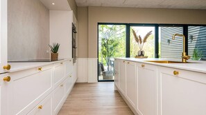 A modern kitchen with white cabinetry and gold handles, a black stove, and a large window with a view of greenery. - VillaVilla 940 - Ebeltoft, Østjylland (Ebeltoft)