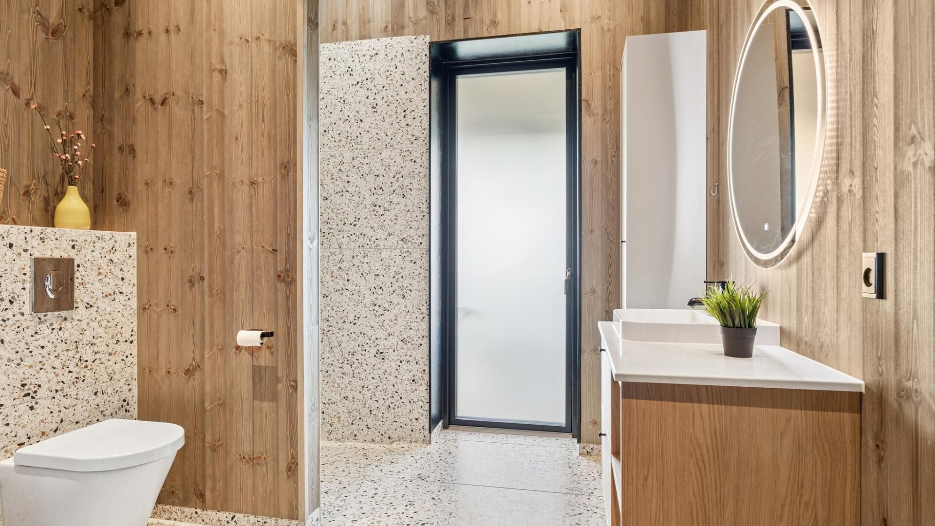 A modern bathroom with a wooden wall, a speckled tile backsplash, a white toilet, a sink with a mirror, and a door leading to another room.