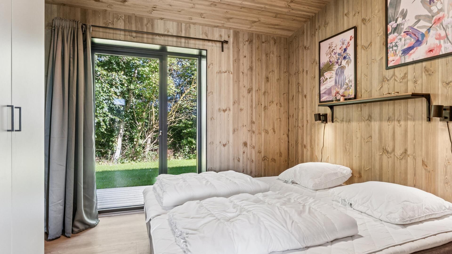 Ein Schlafzimmer mit zwei Betten, einer großen Schiebetür zu einem Garten, Holzwänden und einem gerahmten Kunstwerk über dem Bett.
