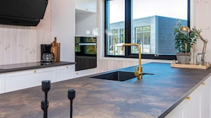A modern kitchen with a black countertop, a gold faucet, and a window overlooking a building. - VillaVilla 938 - Fjellerup, Østjylland (Glesborg)