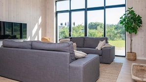 A modern living room with a grey sectional sofa, a flat-screen TV, and a large window offering a view of greenery.
