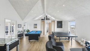 Une pièce spacieuse équipée d’une table de billard, d’une table de ping-pong et d’une table de jeu de fléchettes. Une porte donne accès à un espace extérieur avec une table et des chaises.