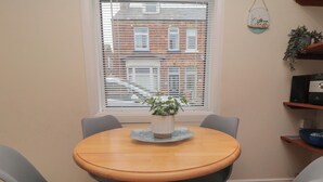 A round wooden table with four grey chairs, a potted plant, and a window with blinds. - Seabreeze (Filey)