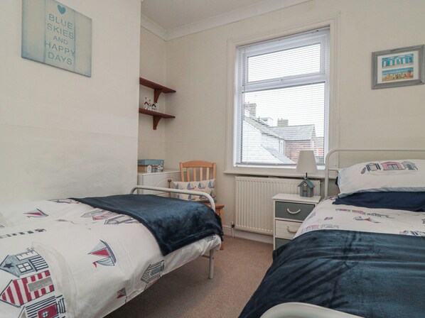 Un dormitorio con dos camas individuales, una ventana con persianas, un radiador, un estante con objetos y una pared con un cuadro enmarcado.