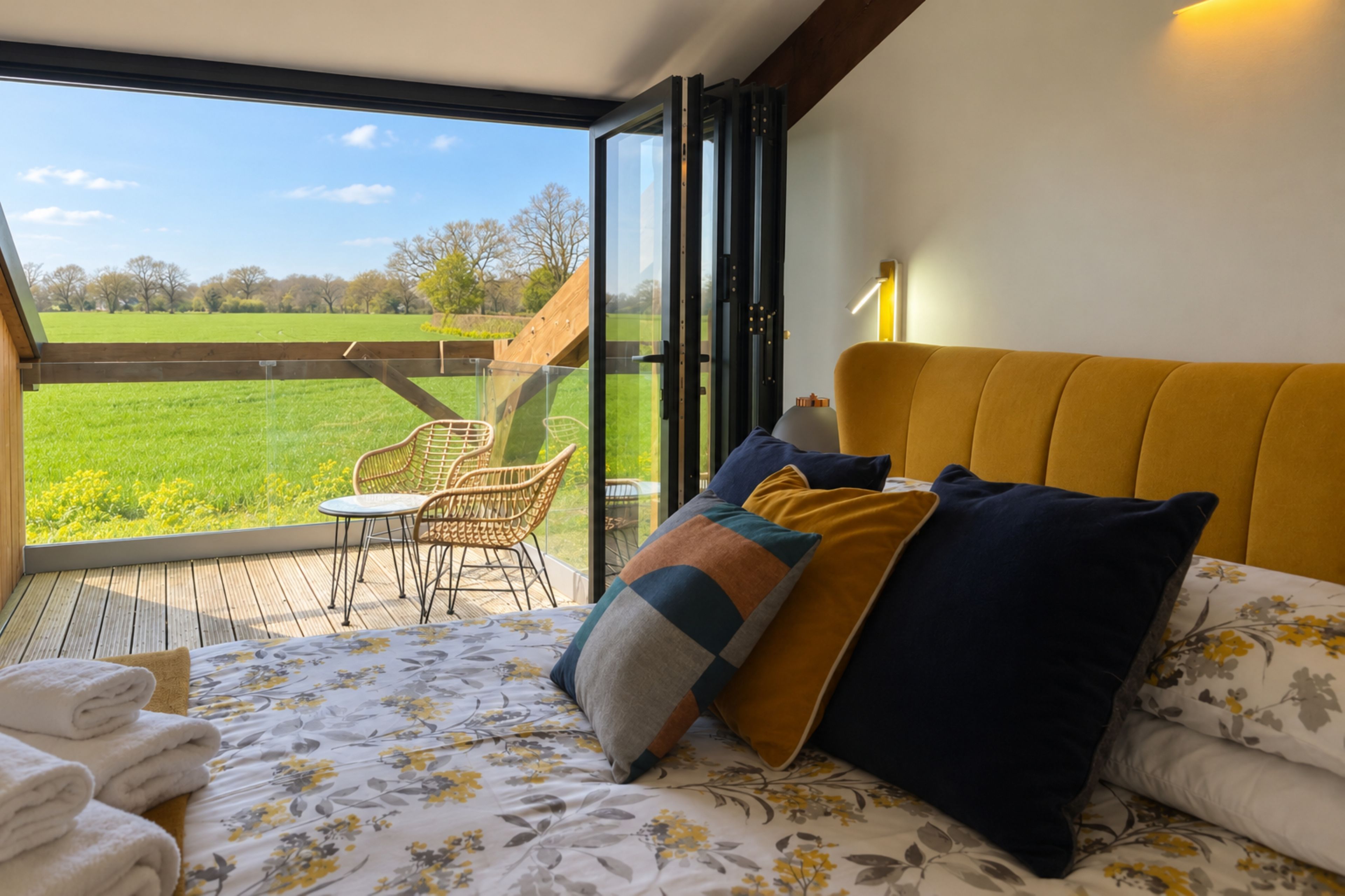 A bedroom with a bed, pillows, a bedside table, a lamp, and a view of a green field through a glass door.
