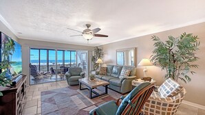 Ein geräumiges Wohnzimmer mit einem großen Fenster, das Blick auf den Strand bietet, einem Deckenventilator und verschiedenen Sitzgelegenheiten, darunter eine Couch und Sessel.