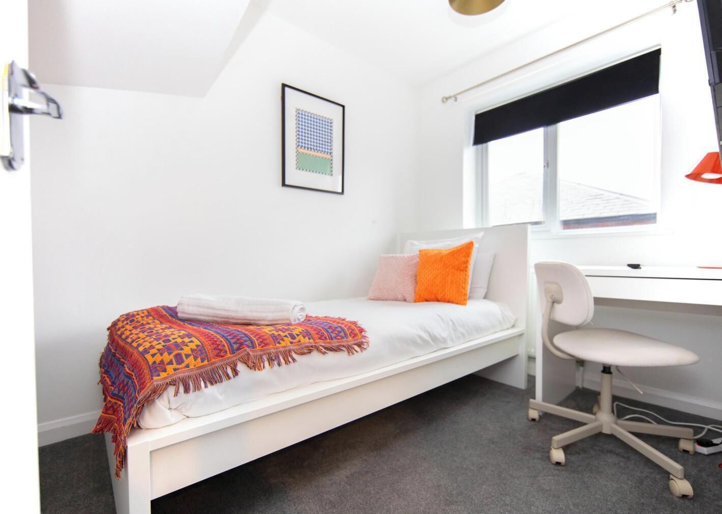 Ein kleines, weißes Zimmer mit einem Einzelbett, einem Schreibtisch und einem Stuhl, einem Fenster mit schwarzem Rollo und einem gerahmten Bild an der Wand.