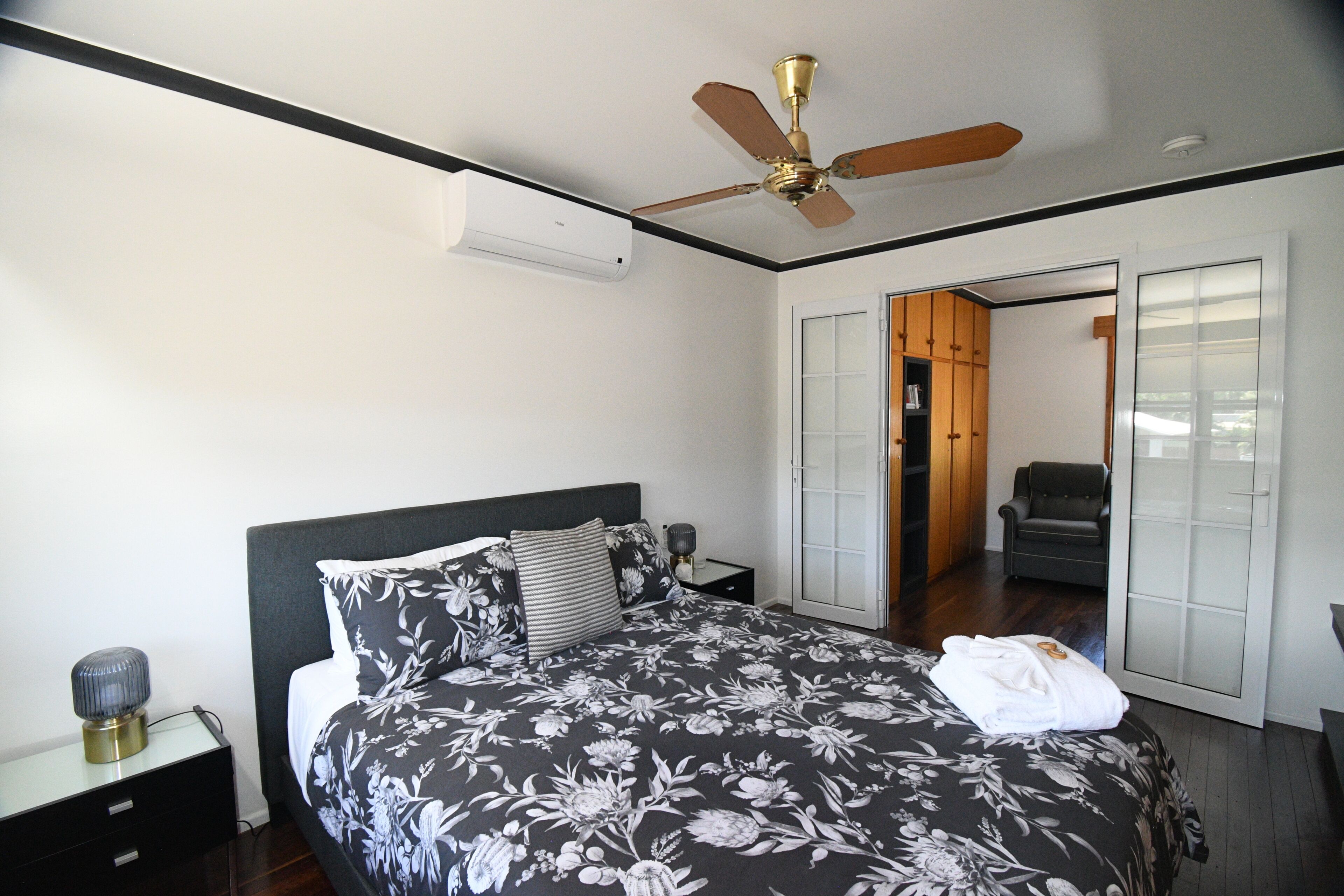 A bedroom with a bed, a ceiling fan, an air conditioning unit, a nightstand, a lamp, and a door leading to another room.