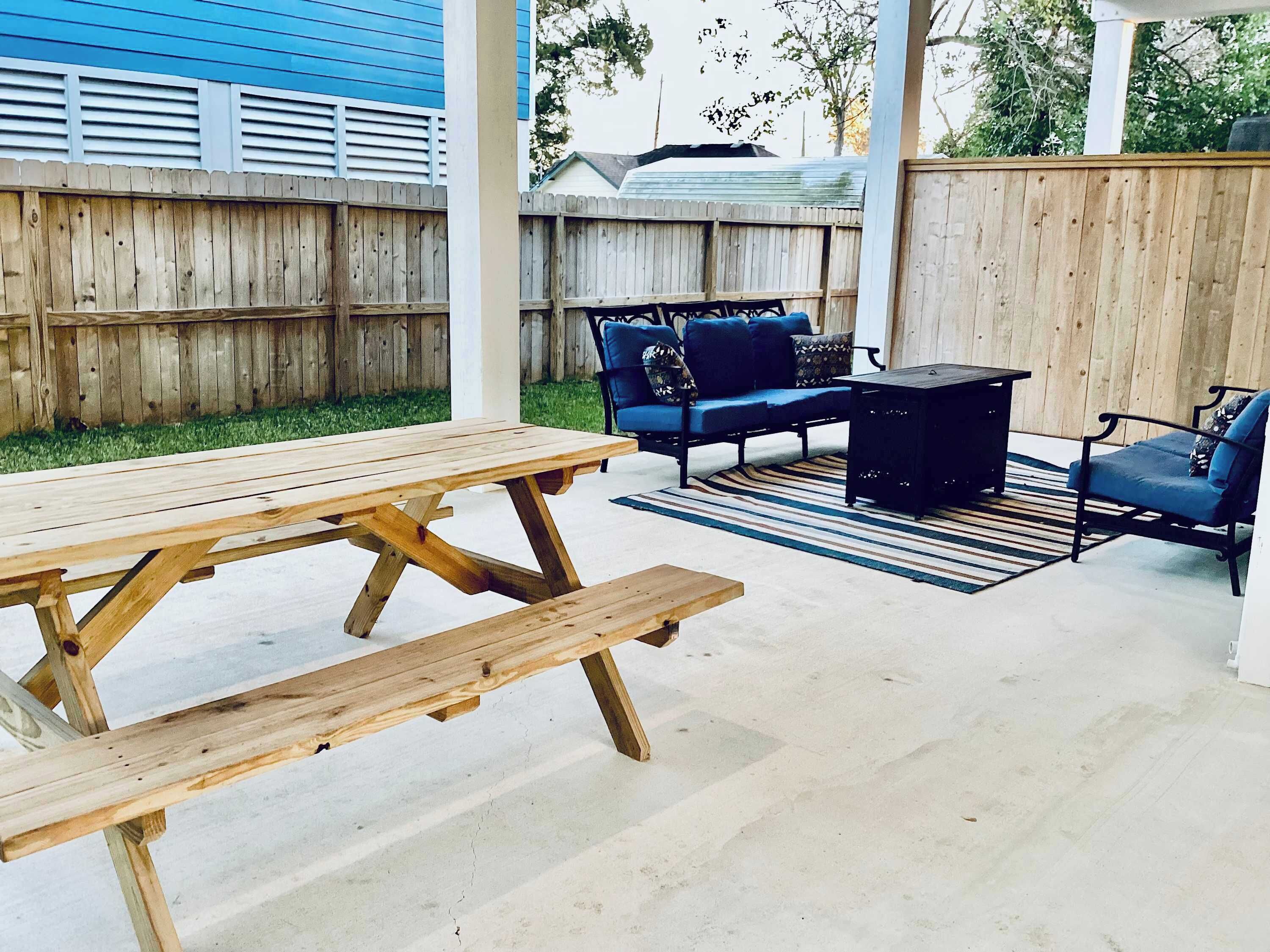 A wooden picnic table and benches in a covered outdoor area with a view of a blue house and a wooden fence in the background.