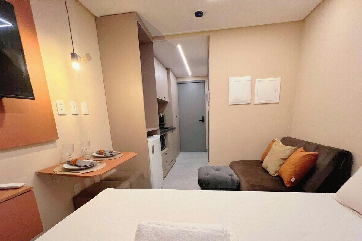 Una habitación de hotel con una cama, un televisor, una mesita con vasos y un plato, un sofá con cojines y un pasillo que conduce a un baño.