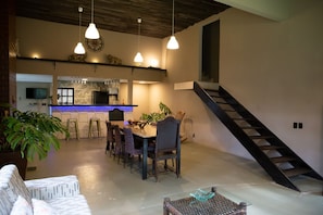 Un vaste espace dédié à la salle à manger, avec une longue table et des chaises, une cuisine en arrière-plan et un escalier menant à un niveau supérieur.