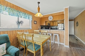 A dining area with a wooden table and chairs, a kitchen with wooden cabinets and appliances, and a window with blinds. - Mariner's Cove 110A- Marsh View 2BR- Pool (Myrtle Beach)