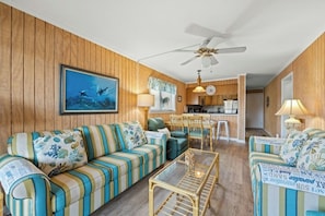 A living room with striped sofas, a glass coffee table, and a ceiling fan. - Mariner's Cove 110A- Marsh View 2BR- Pool (Myrtle Beach)