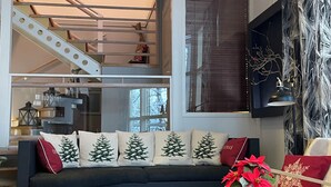 Ein modernes Wohnzimmer mit einem dunkelgrauen Sofa, weihnachtlichen Kissen, einem Couchtisch mit dekorativer Mitte und einer Treppe, die in eine obere Etage führt.