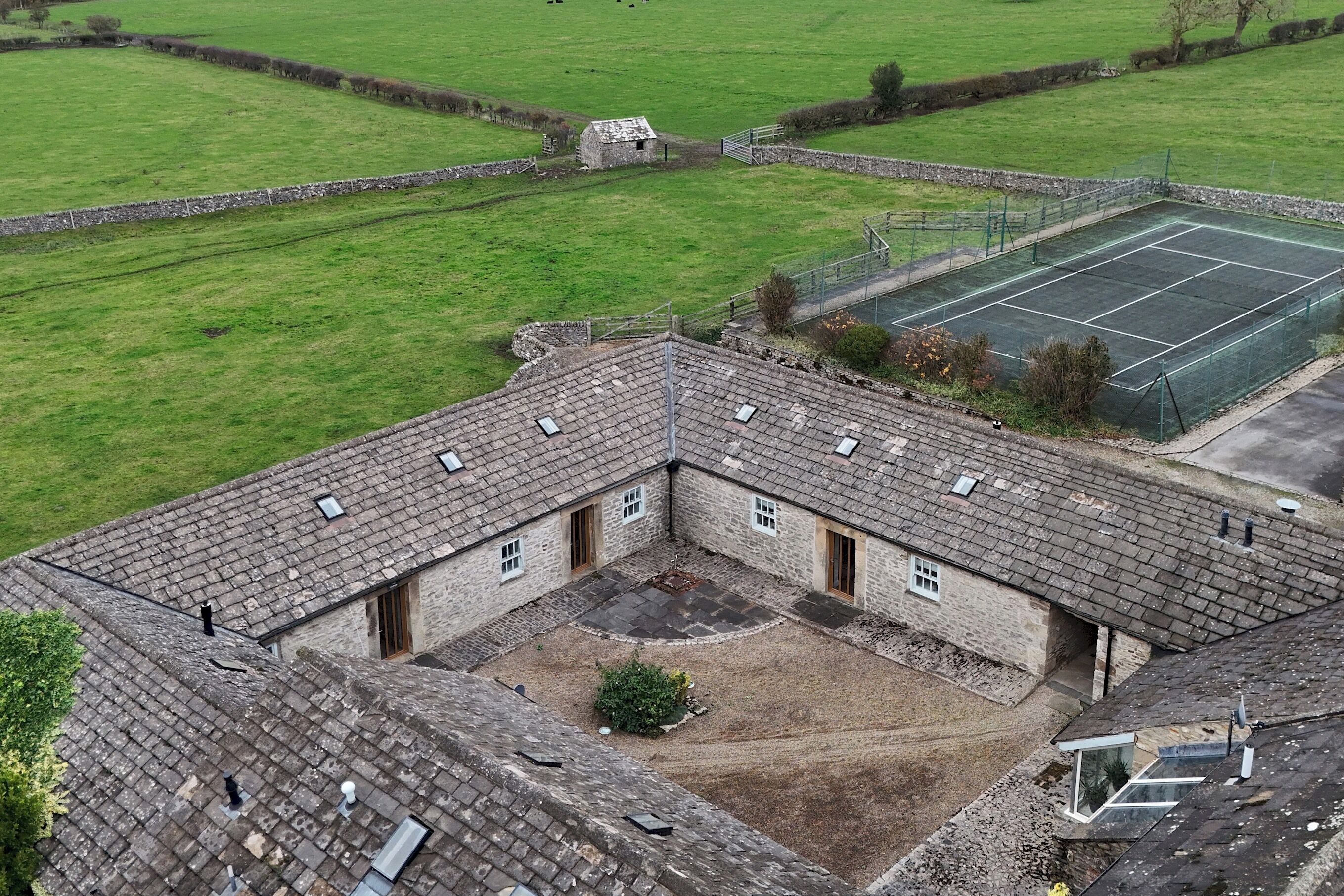 A courtyard with a tennis court, stone buildings, and a grassy field.